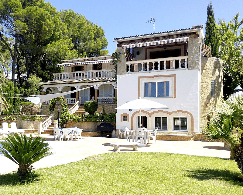 Fachada posterior de una villa blanca de varios niveles con detalles en piedra, piscina, palmeras y terrazas.