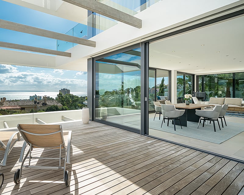 Sun loungers on a wooden terrace with glass railings overlooking the sea, adjacent to an open-plan dining room.