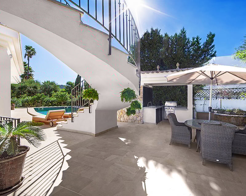 Sunny terrace with outdoor kitchen, wicker dining set under a parasol, and a white spiral staircase near a pool.