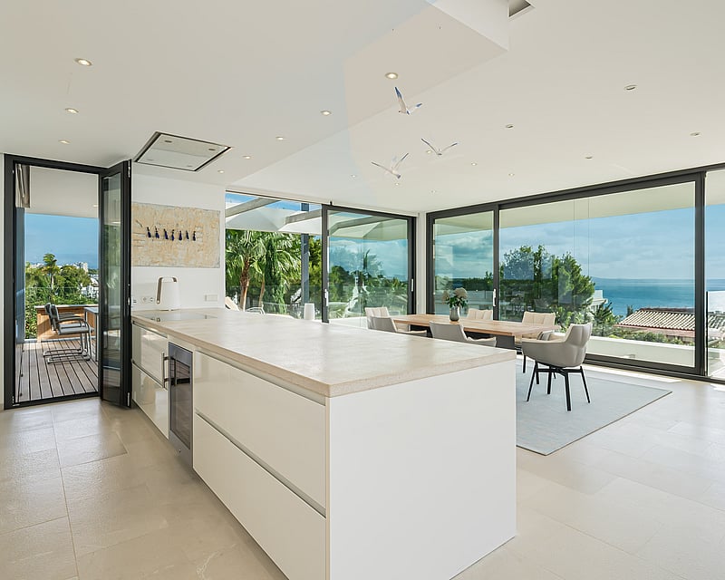 Bright modern white kitchen with large island, dining area, and floor-to-ceiling glass walls overlooking the ocean.