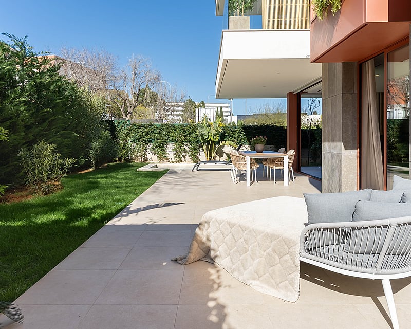 Modern terrace with stone tiling, outdoor dining set, designer lounge chair, and a manicured green lawn under a blue sky.