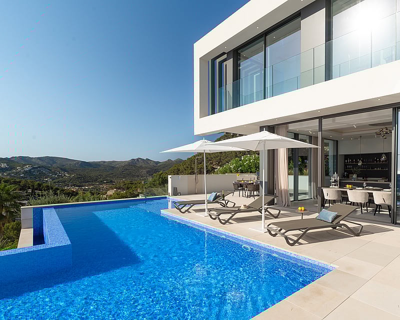 Luxury infinity pool and white modern villa with mountain views and designer loungers under white umbrellas.