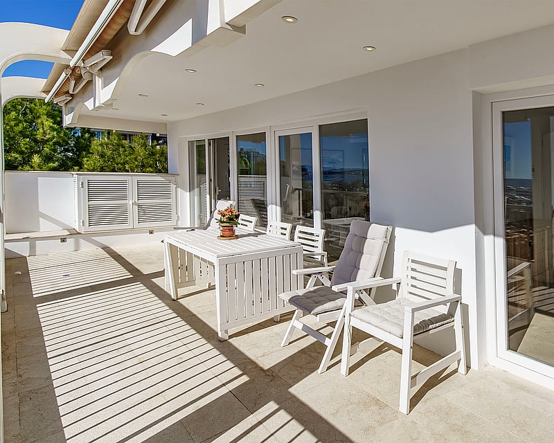 Sunlit terrace with white outdoor dining set and large glass sliding doors reflecting the coastline.
