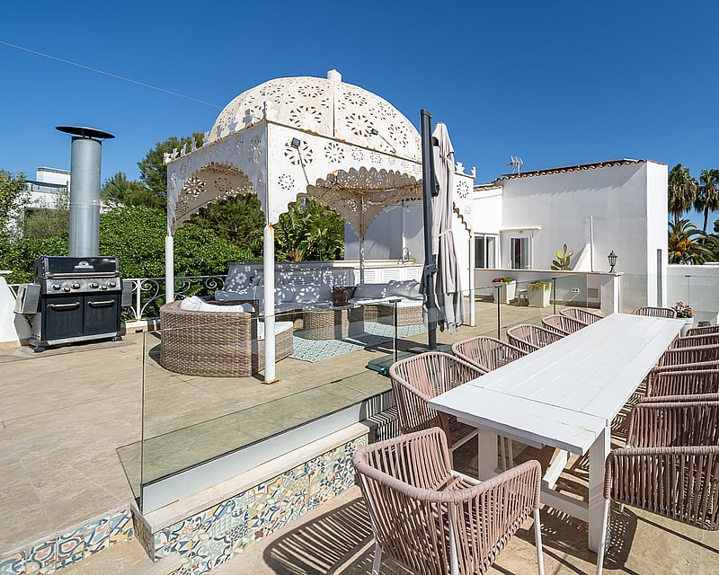 Spacious terrace with a white dining table, wicker chairs, BBQ station, and an ornate white gazebo under a clear blue sky.