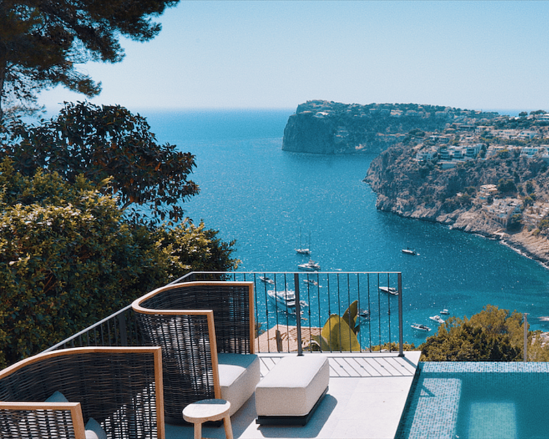 Luxury terrace featuring an infinity pool and designer lounge chairs overlooking a bay with yachts and cliffs.