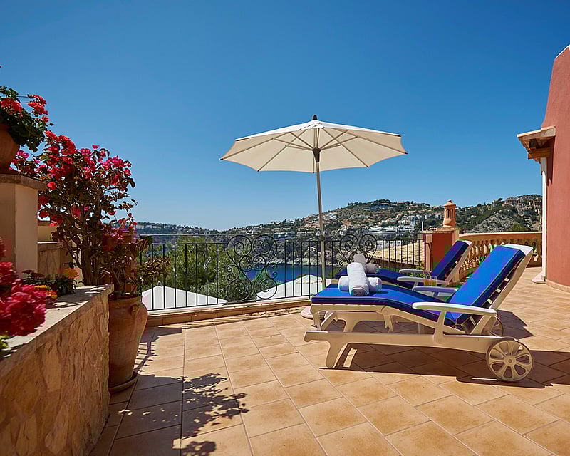 Sunny terrace with two blue lounge chairs, white umbrella, and panoramic sea and coastal hillside views.