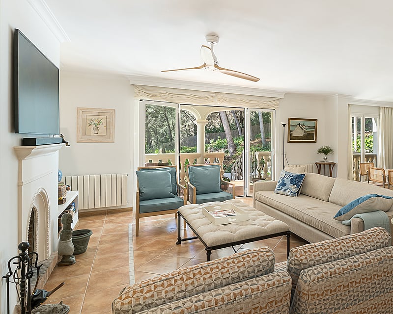 Spacious living room with terracotta floors, cream sofas, fireplace, and glass doors opening to a lush garden.