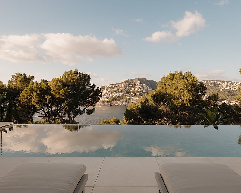 Infinity pool overlooking a Mediterranean bay and forested hills with sun loungers in the foreground.