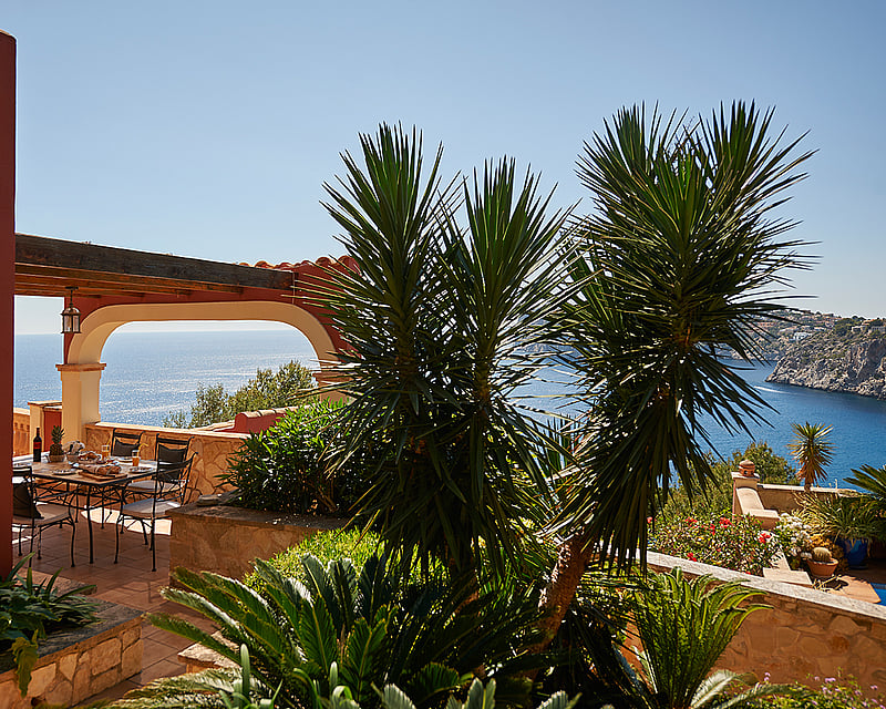 Outdoor dining table on a terracotta terrace with an arched pergola overlooking the blue sea and lush greenery.