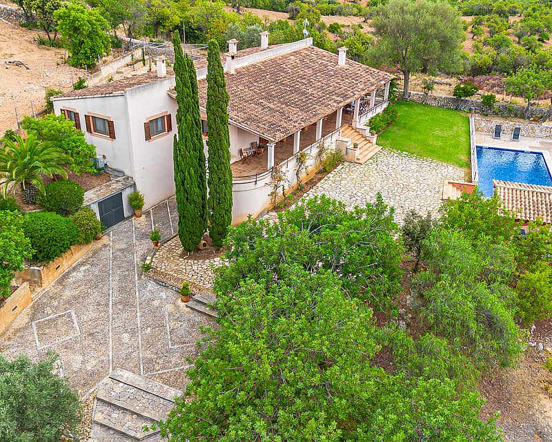 High-angle view of a villa with a terracotta roof, stone courtyard, blue swimming pool, and lush green landscaping.