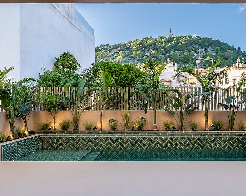 A modern rectangular pool with green herringbone tiles, surrounded by palm trees and plants under a blue sky.