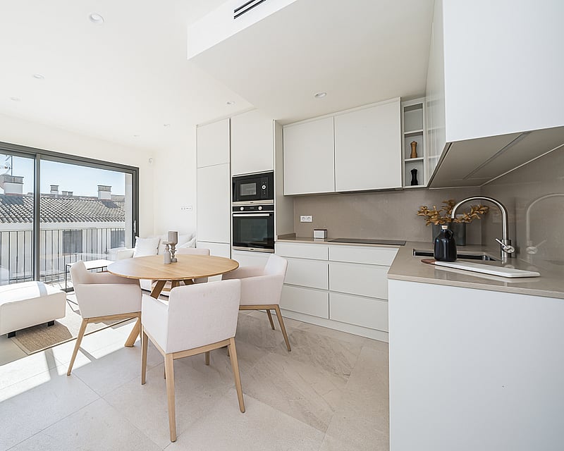 Minimalist white kitchen featuring an oak dining table, upholstered chairs, and large sliding glass doors.