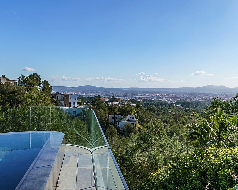 An infinity-edge pool with glass railing overlooking a lush forested valley and distant city.