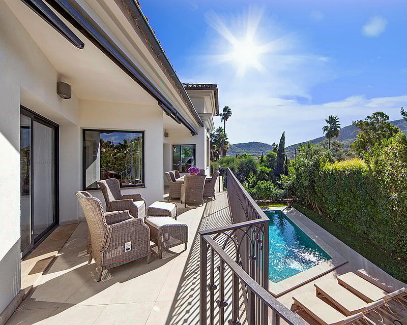 Sunny elevated terrace with wicker lounge furniture overlooking a private pool, green gardens, and distant mountains.