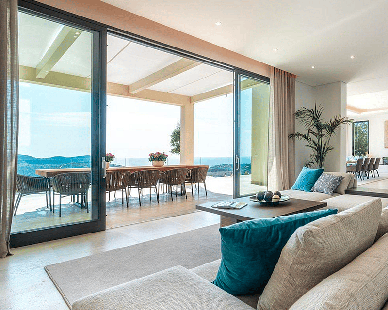 Spacious modern living room with large beige sofa, floor-to-ceiling glass doors opening to a terrace with sea views.