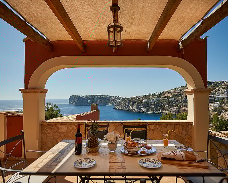 A covered Mediterranean terrace with a breakfast-set table looking out over a blue bay and coastal cliffs under a clear sky.