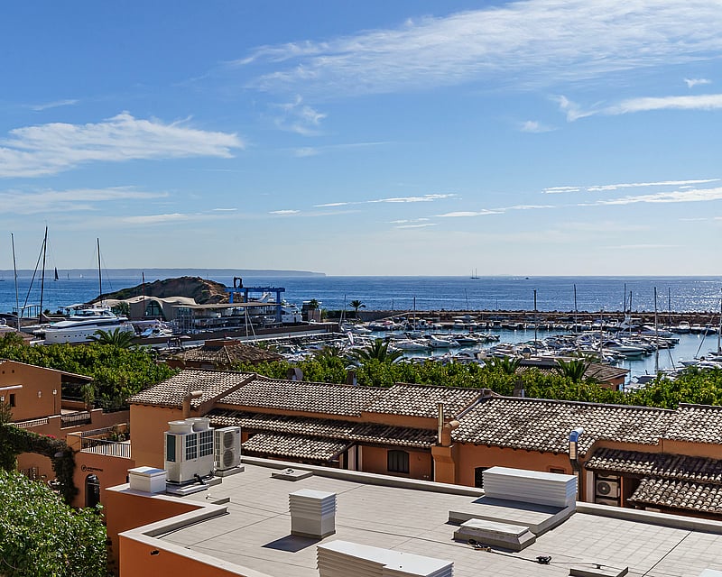 Elevated view of a Mediterranean marina with terracotta roofs, white yachts, and the blue sea under a clear sky.