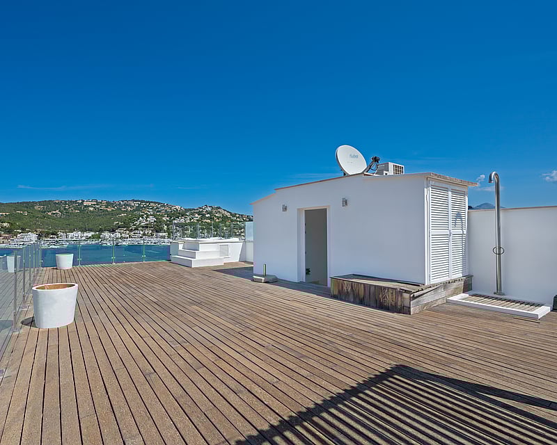 Spacious wooden rooftop terrace with glass railings overlooking a Mediterranean marina and green hills.
