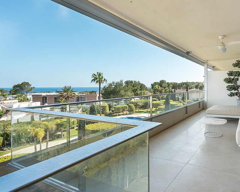 Wide modern terrace with white outdoor sofas, glass railings, and views of lush gardens and the sea in the distance.