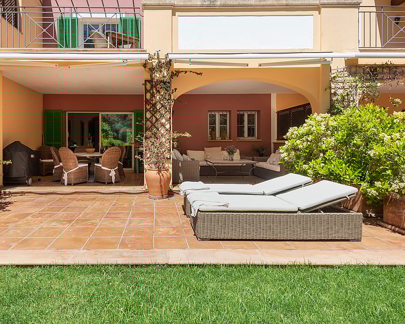 A sunny terracotta terrace with wicker lounge furniture and sunbeds bordering a lush green lawn.