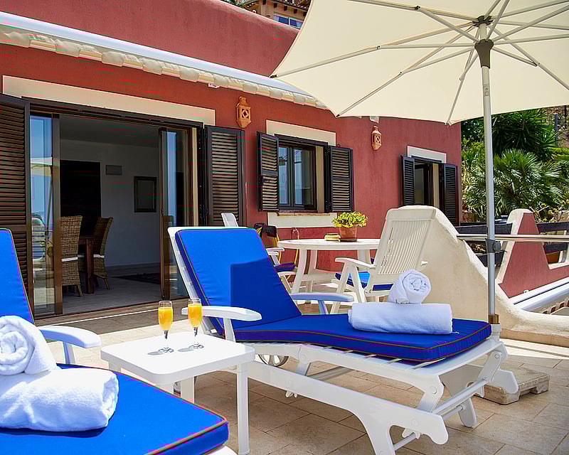 Private terrace with blue sun loungers, white towels, orange juice, and an umbrella against a terracotta building.