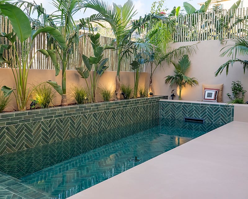 A luxury swimming pool with green herringbone tiles, surrounded by palm trees and beige walls with warm spotlighting.