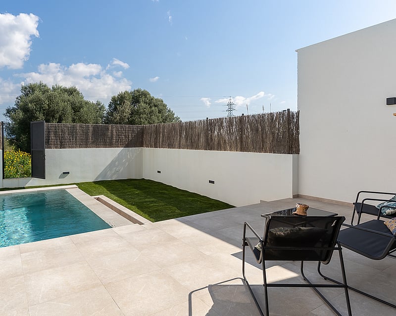 Private swimming pool with light stone tiling, a small lawn area, and reed fencing under a bright blue sky.