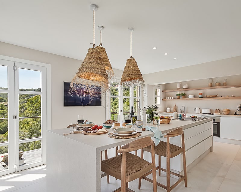 Bright modern kitchen featuring a large white island, wicker pendant lights, and floor-to-ceiling windows with forest views.