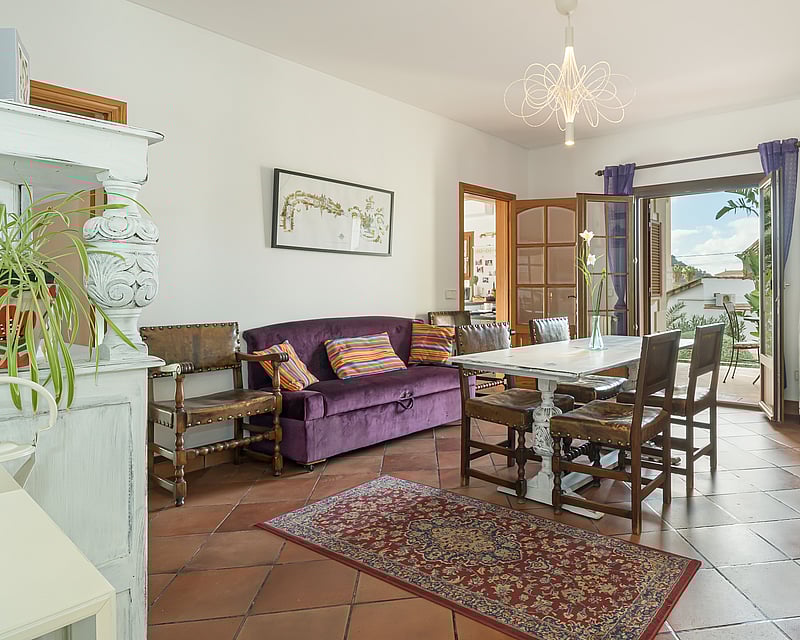 Dining room with terracotta floors, purple velvet sofa, and white wooden table opening to a sunny balcony.