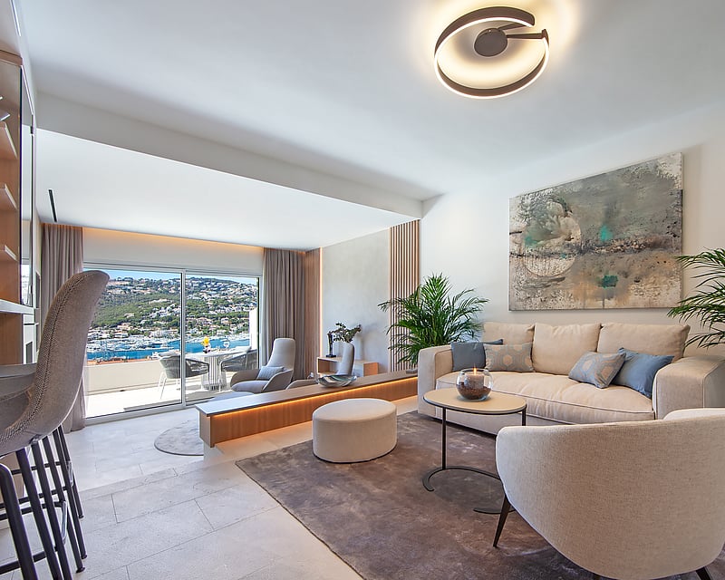 Bright modern living room with cream furniture, large windows overlooking the sea, and lush indoor plants.