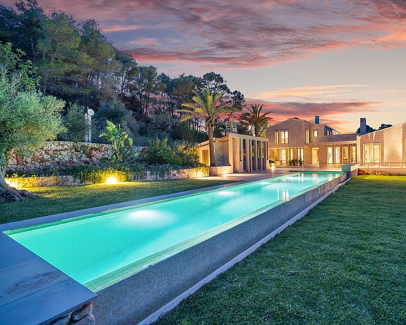 Long glowing lap pool leading to a modern stone villa under a vibrant orange and pink sunset sky.