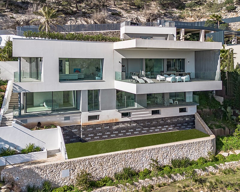 Modern white multi-story villa with large glass windows and terraces built into a rocky hillside with stone walls.