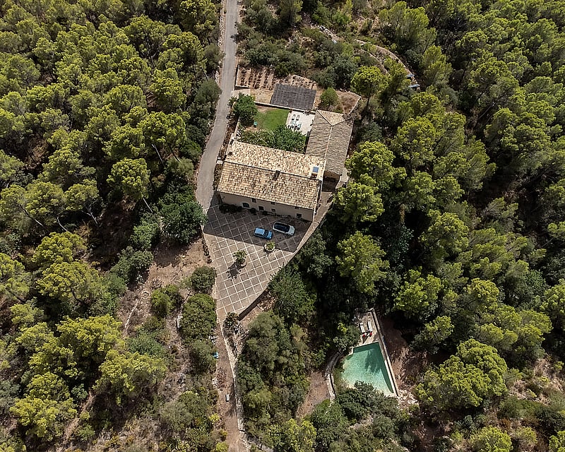 Aerial view of a stone house with terracotta roof, courtyard, and a pool, all surrounded by a dense green pine forest.
