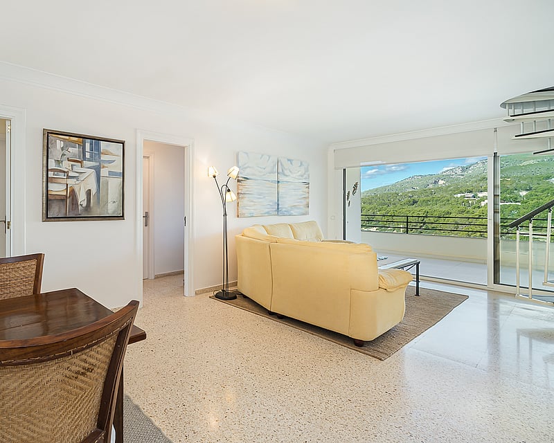 Open living space with a cream leather sofa, wooden dining set, and large glass doors revealing a mountain landscape.