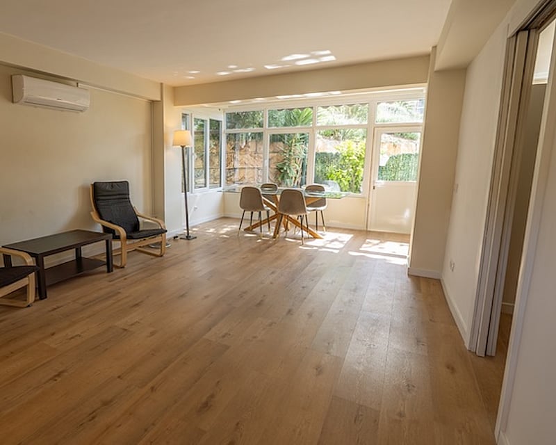 Salón con suelos de madera, grandes ventanales con vistas al jardín, mesa de comedor de cristal y dos sillones negros.