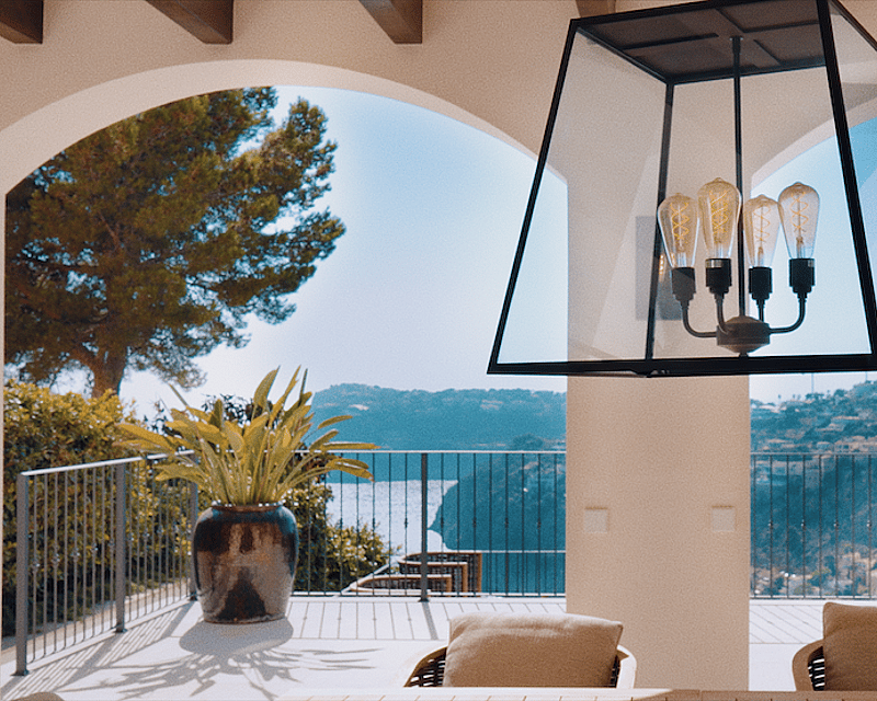 View through white arches from a villa terrace overlooking the sea and green hills under a bright blue sky.