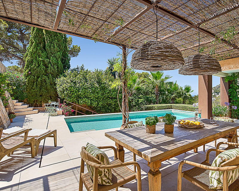 Outdoor dining area under a reed pergola next to a swimming pool with mountain views and lush greenery.