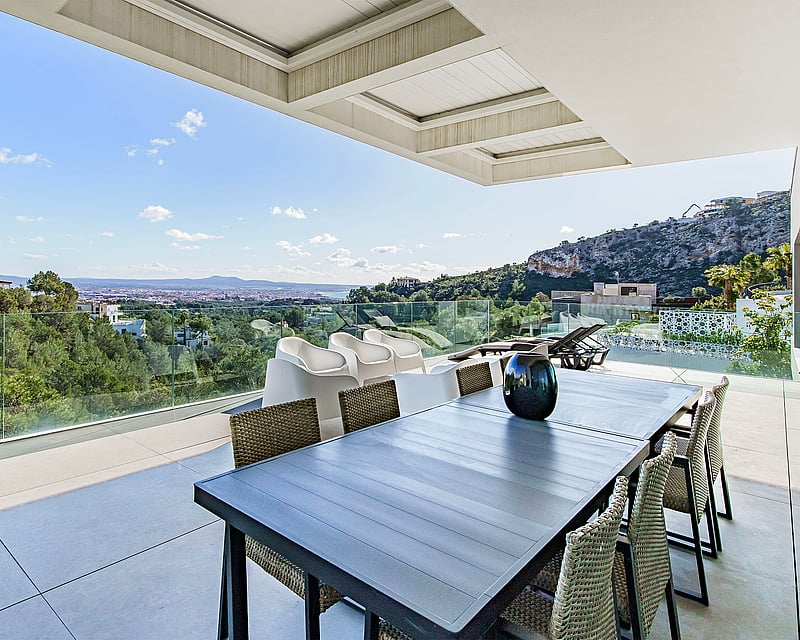 Modern terrace with a dining table and glass railings overlooking a lush green forest and distant city.