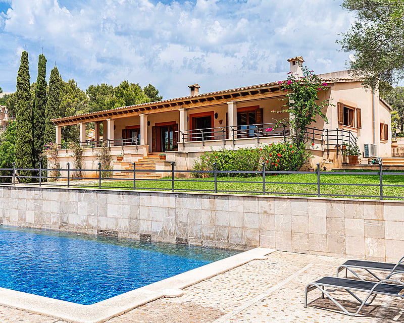 Large blue swimming pool and sun loungers in front of a Mediterranean villa with a stone wall, lawn, and covered terrace.