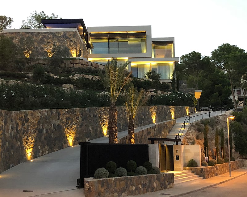 Architectural evening shot of a tiered luxury villa with stone walls, glass balconies, and illuminated driveway.