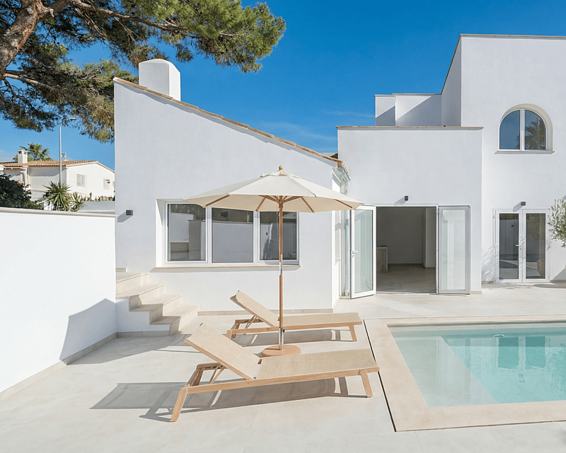 Contemporary white villa featuring a rectangular swimming pool, light stone terrace, and sun loungers under a clear blue sky.