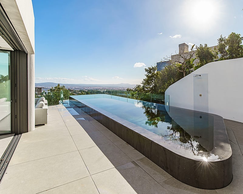 A sleek, long infinity pool on a minimalist white terrace overlooking a distant city under a bright blue sky.