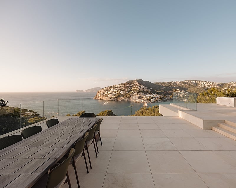Elevated stone terrace with a wooden dining table overlooking a Mediterranean bay and coastal mountains at sunset.