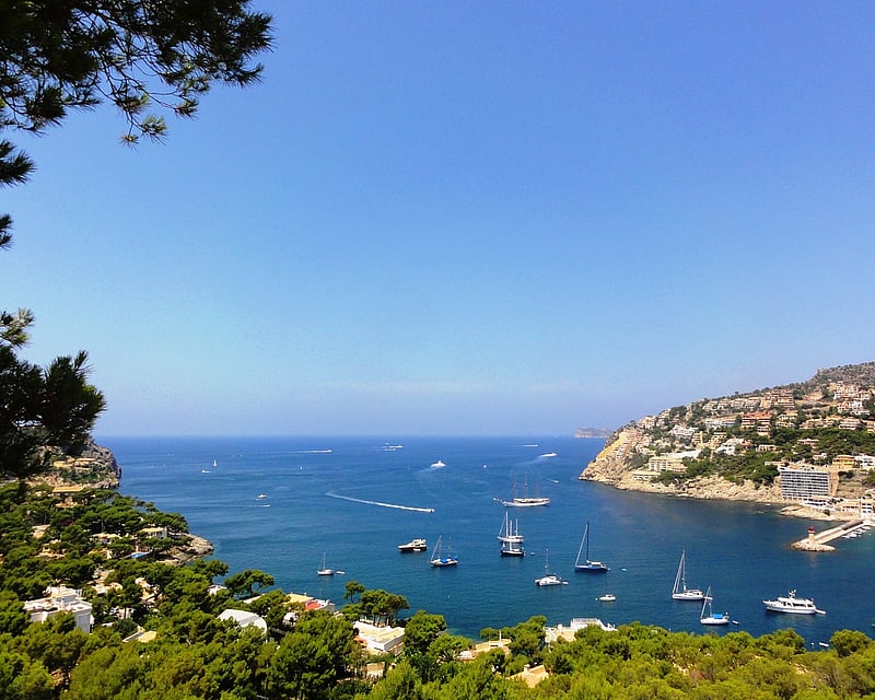 High-angle view of a blue bay with sailboats, surrounded by green hills and Mediterranean villas under a clear sky.