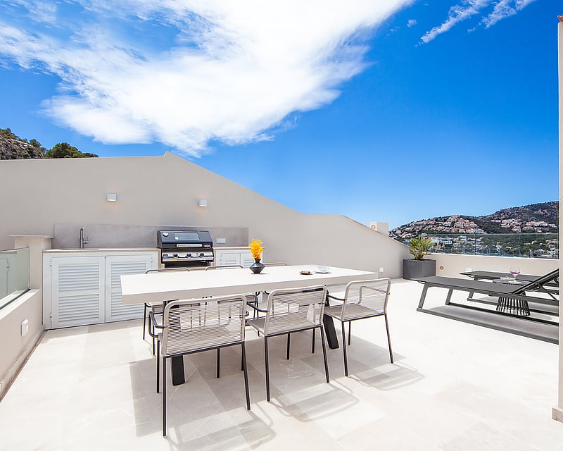 Modern rooftop terrace featuring an outdoor kitchen, dining table, and sun loungers with panoramic coastal views.