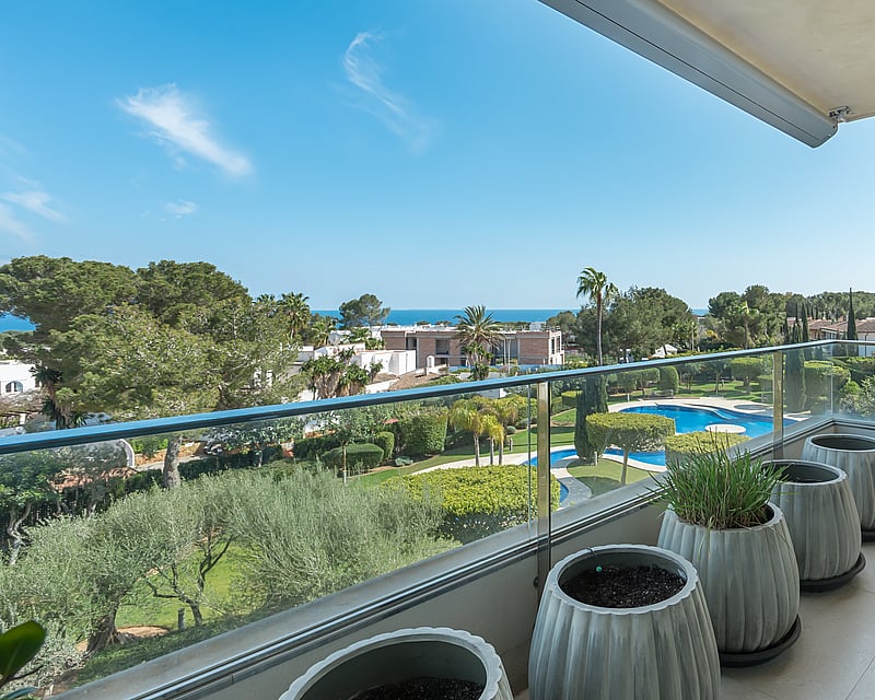 Modern balcony with glass railings overlooking lush green gardens and the blue sea under a clear sky.