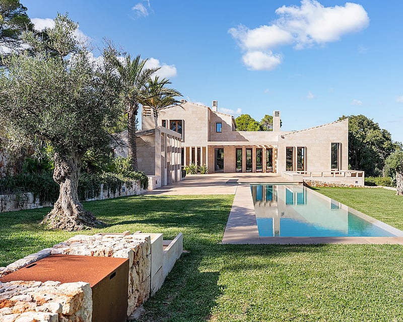 Modern sandstone villa with a long rectangular lap pool, lush green lawn, and ancient olive trees under a blue sky.