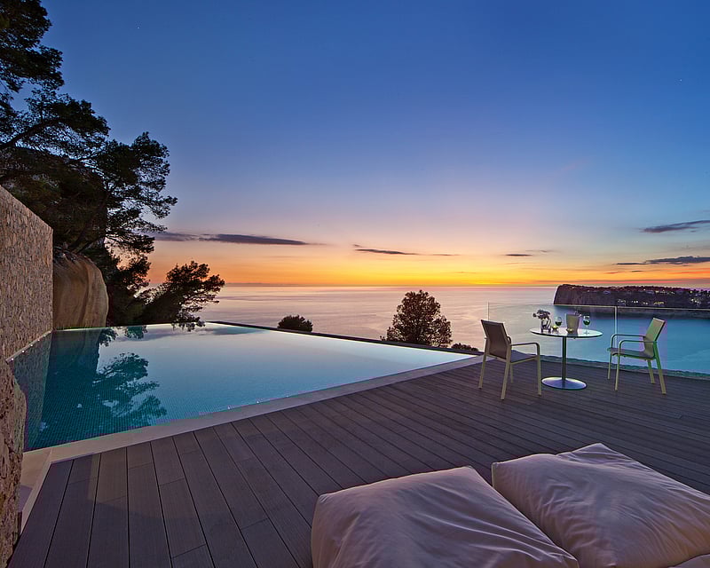 Infinity pool on a wooden deck overlooking the sea at sunset with outdoor furniture and glass railings.