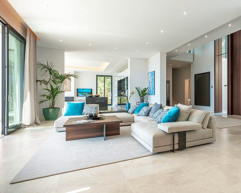 Bright living room with a large beige sectional sofa, turquoise pillows, marble floors, and floor-to-ceiling windows.