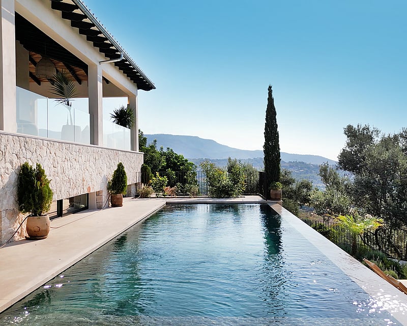 An infinity edge swimming pool next to a stone-walled villa with glass railings, overlooking a mountain valley.